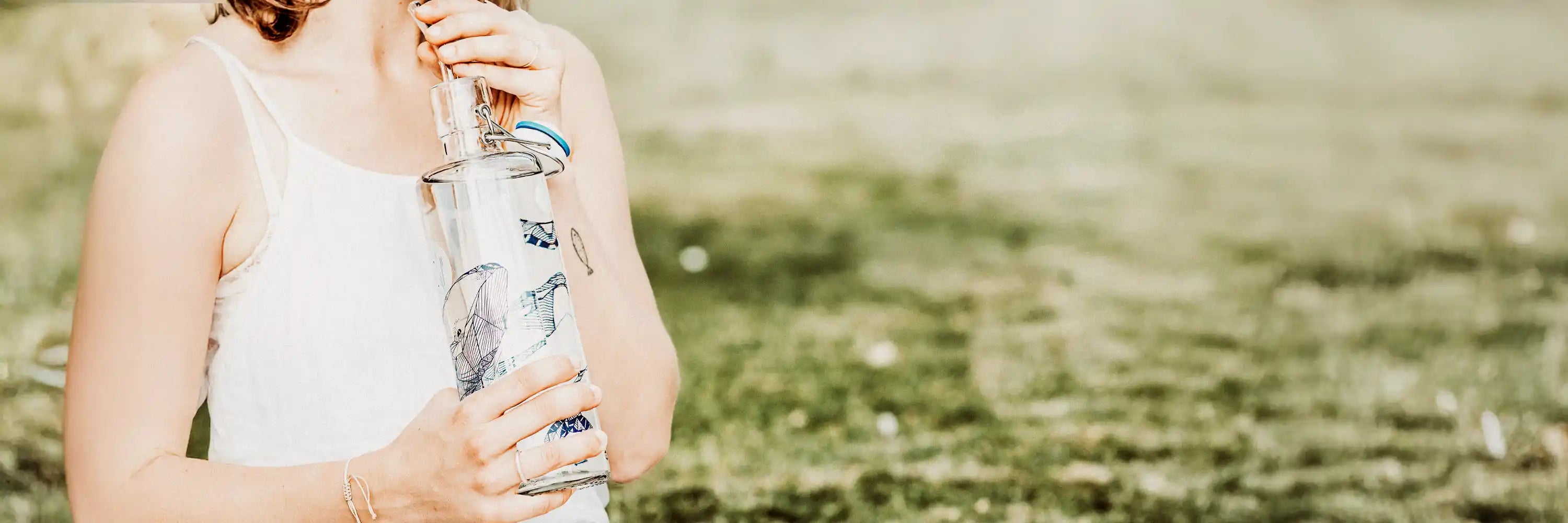 Frau mit weißem Top und Trinkflasche aus Glas in der Hand auf wiese im Park